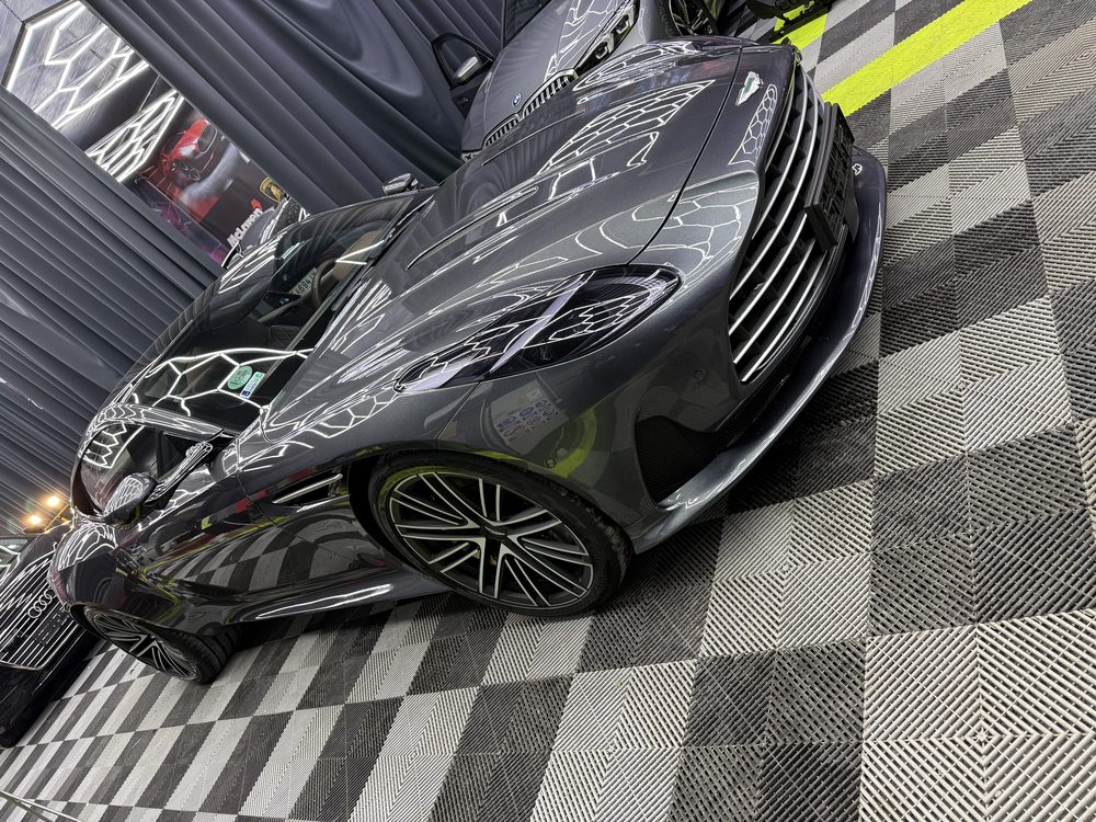 Aston Martin gri-antracit văzut de aproape din față-stânga, cu luminile hexagonale ale atelierului oglindite pe capotă