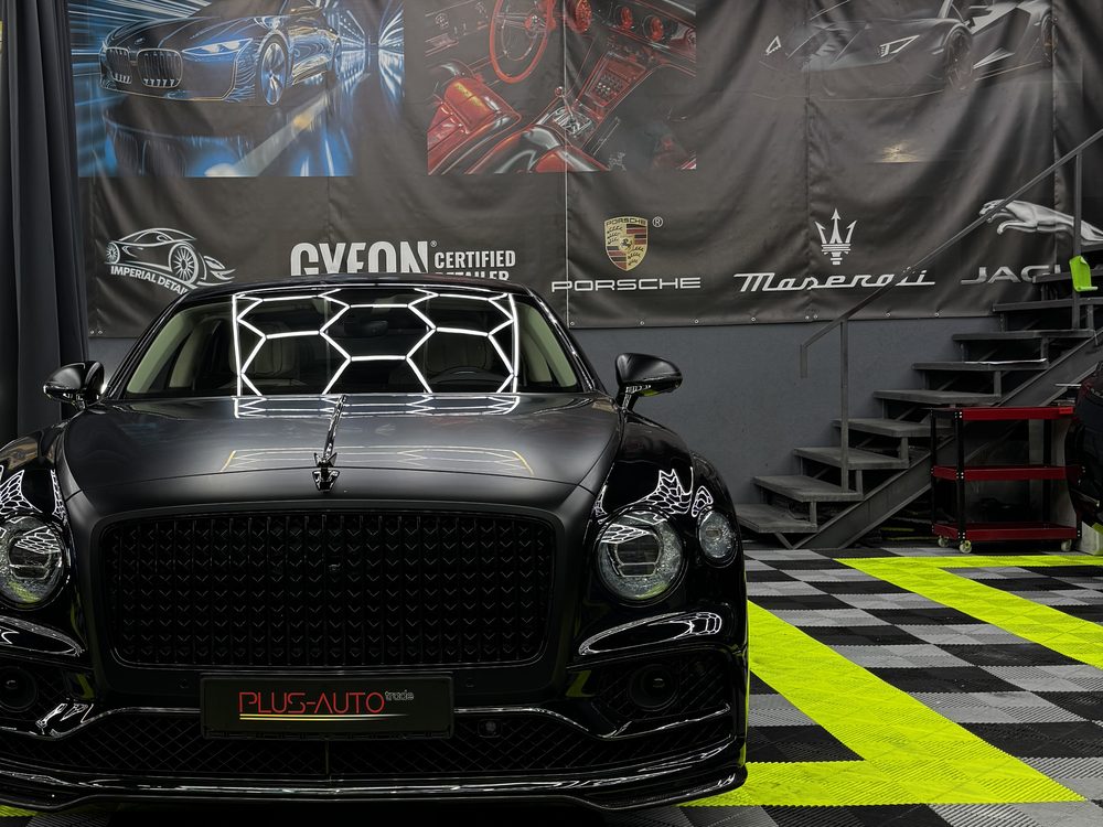 Bentley Flying Spur negru mat, văzut din față, cu luminile hexagonale ale atelierului deasupra și podea în carouri