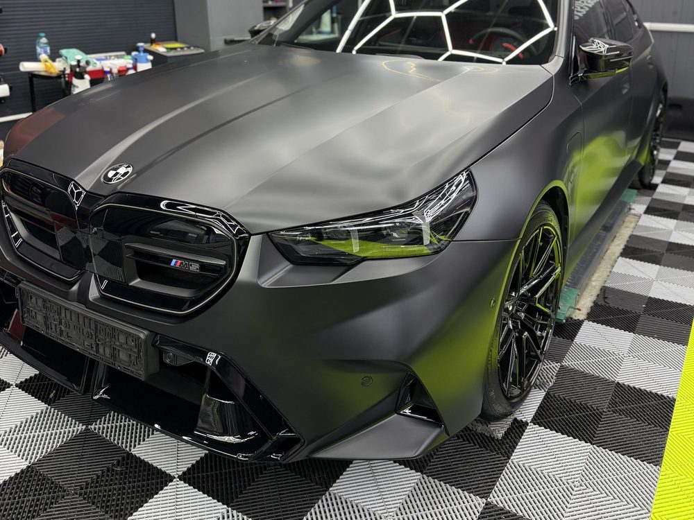 BMW M5 gri închis satinat, văzut din față-stânga, cu grilă neagră lucioasă și jante negre