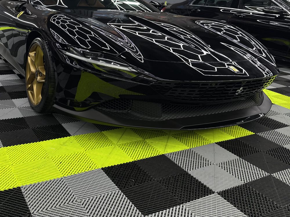Ferrari negru cu jante aurii, văzut din față-stânga, cu luminile hexagonale ale atelierului oglindite pe capota lungă