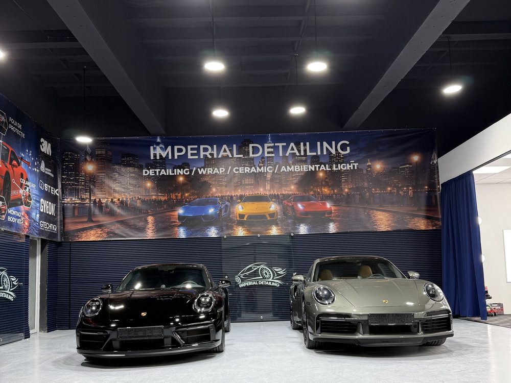 Două Porsche 911, unul negru și unul verde-măsliniu, parcate unul lângă altul pe sigla Imperial Detailing Oradea de pe pardoseală