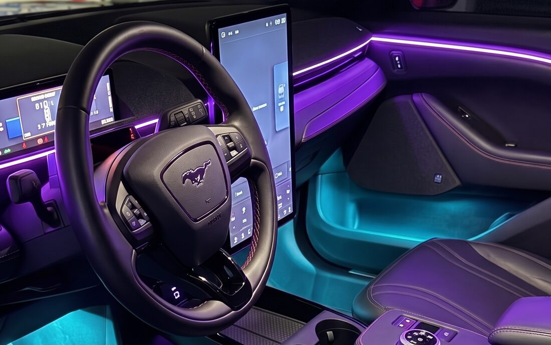 Același interior de Ford Mustang Mach-E, cu iluminarea ambientală aprinsă: linie mov pe bord și portieră și lumină turcoaz în zona picioarelor
